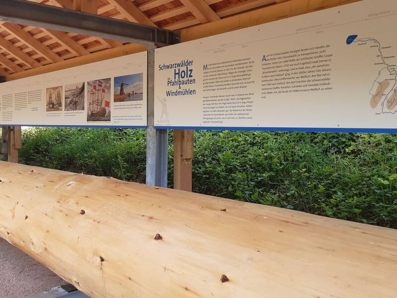 liegender Holländerstamm zum Anfassen und Draufsitzen unter einem Holzdach mit Infotafeln.