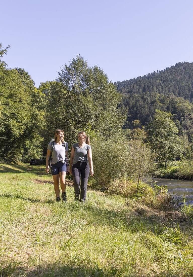 Zwei Wanderinnen gehen über eine sonnige Wiese am Ufer der Kinzig mit bewaldeten Höhenrücken im Hintergrund