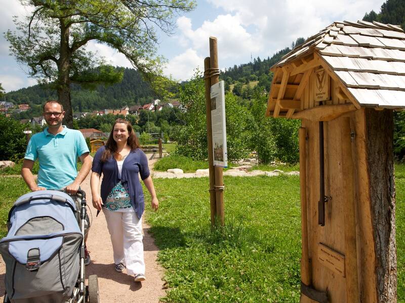 Eine Familie mit Kinderwagen läuft durch einen Park neben einer Infotafel zum Flößerpfad und ausgestellten Flößerwerkzeugen
